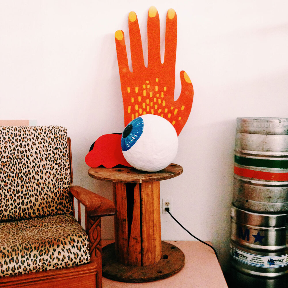 A comically sized orange hand and an eyeball larger than a basketball sit on top of a wood wire spool repurpossed as a cocktail table. Empty beer kegs are stacked on on the floor and a leopard print couch is next to the table.